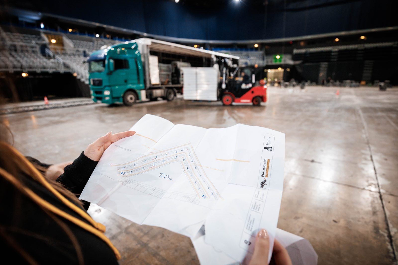 Gefalteter Bestuhlungsplan in der Hand, im Hintergrund die Anlieferung von Bühnenelementen mit Lkw und Gabelstapler in der Halle