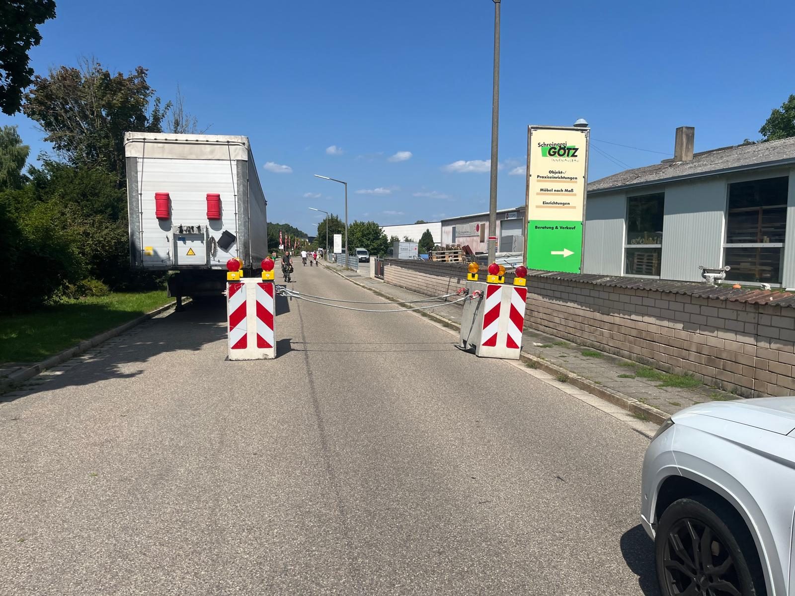 Gesperrte Zufahrtsstraße mit zwei rot-weißen Absperrbaken und gespanntem Stahlseil, links ein Sattelauflieger