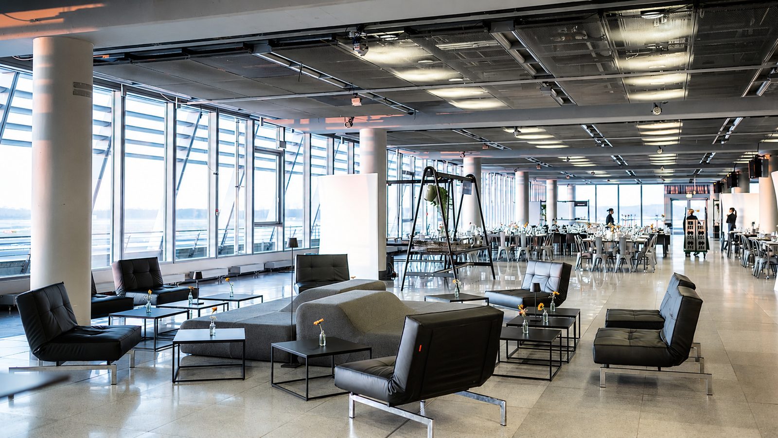 Lounge-Bestuhlung mit schwarzen Ledersesseln und Sofa in einer Halle mit Glasfassade, dahinter eingedeckte Tische