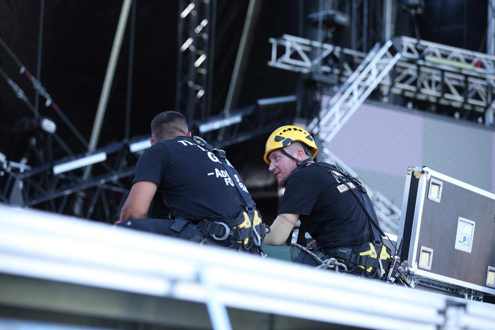Zwei Rigger mit Auffanggurt stimmen sich auf einer Bühnenkonstruktion vor Aluminium-Traversen ab, der rechte trägt einen gelben Kletterhelm