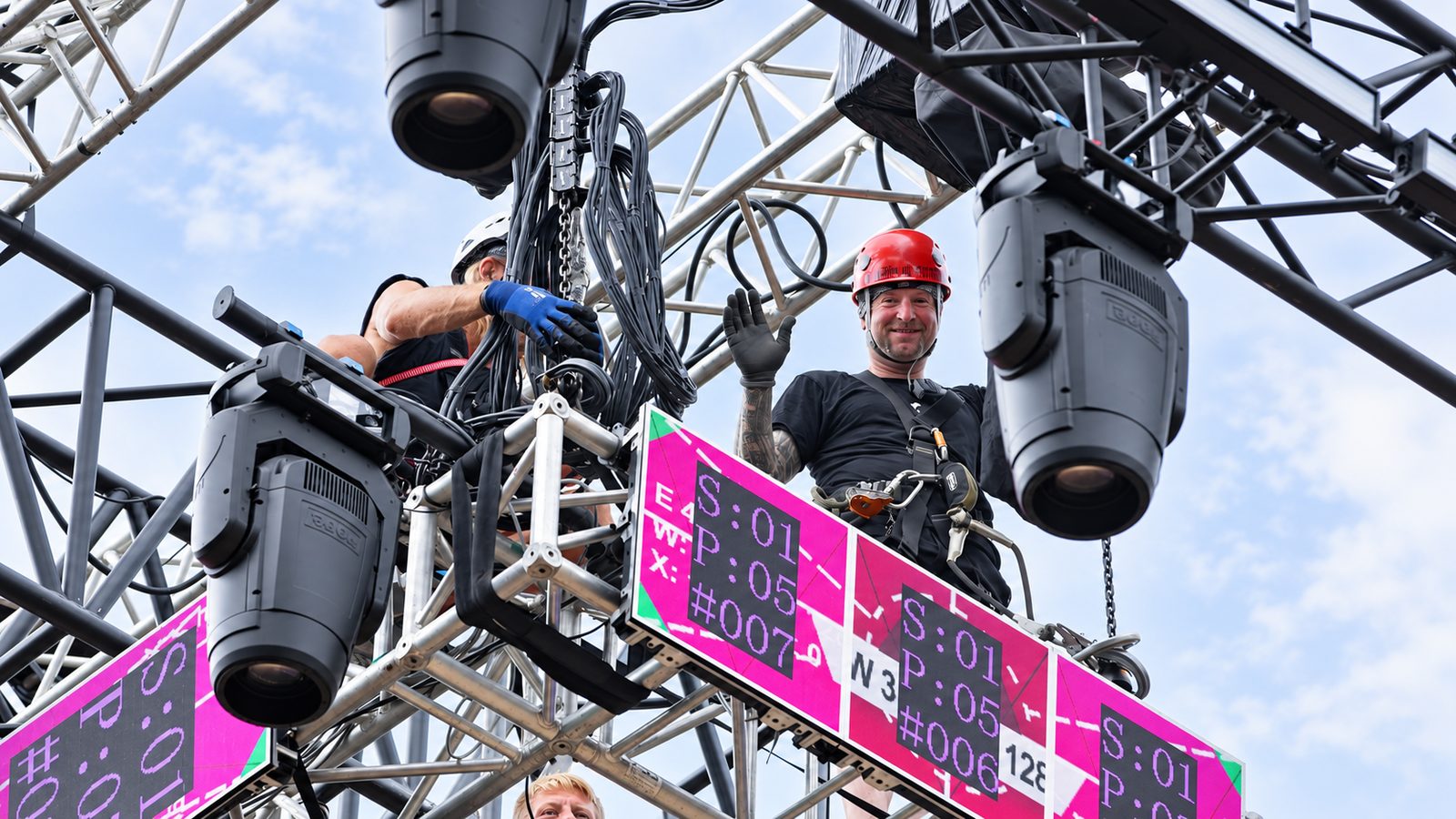 Techniker mit rotem und weißem Schutzhelm arbeiten auf einer Traverse an Scheinwerfern und Kabeln
