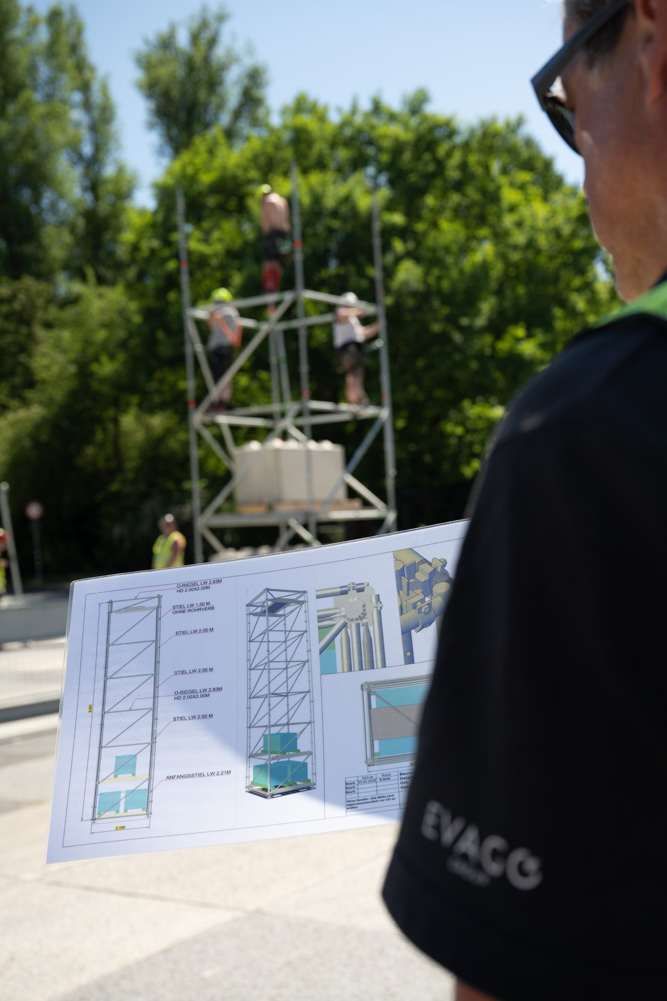 Eventproduktion: Aufbauplan eines Gerüstturms in der Hand, dahinter errichtet ein Team dieselbe Konstruktion
