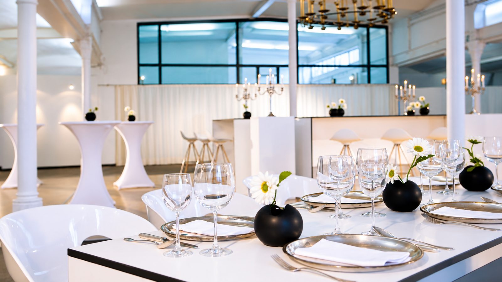 Gedeckte Bankettafel mit Silberplatztellern und Weingläsern in einer weißen Halle mit Stehtischen
