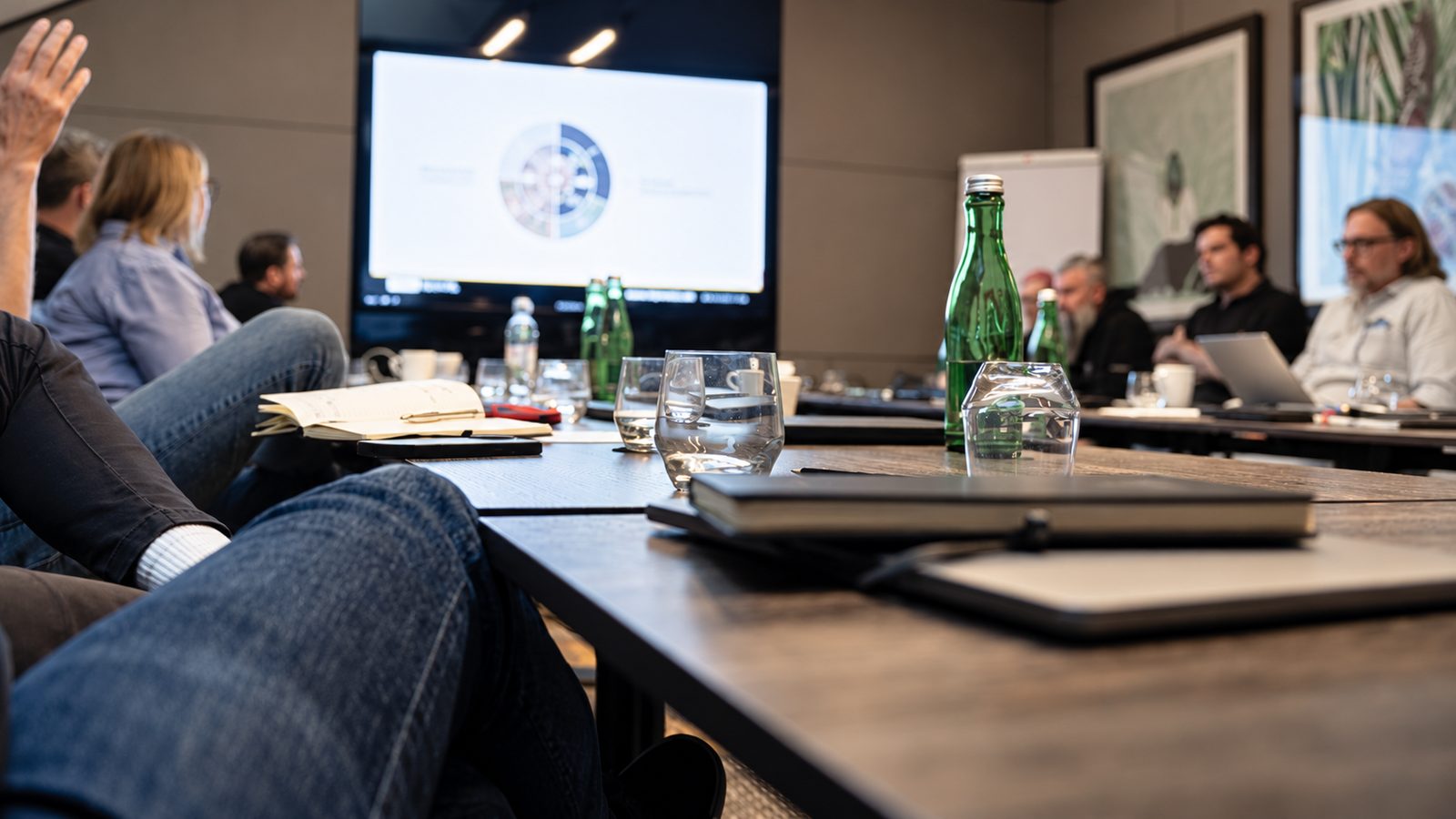 Teilnehmer im Boardroom, eine erhobene Hand zur Wortmeldung, im Hintergrund eine Folie mit Kreisdiagramm