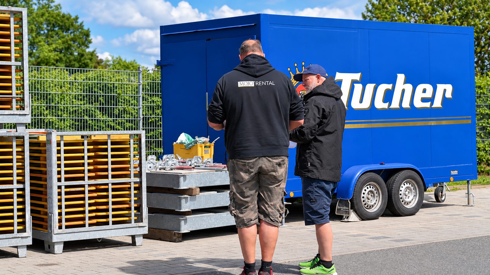 Zwei Mitarbeiter besprechen Unterlagen zwischen Gitterboxen mit gelben Stangen und einem Anhänger