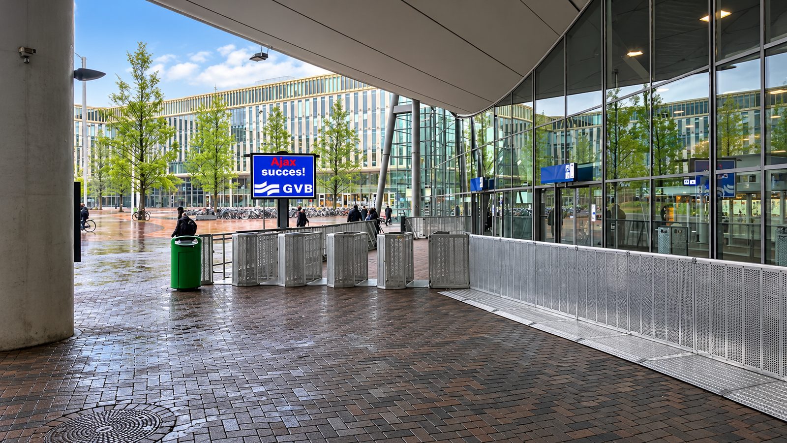 Reihe silberner Drehkreuze und perforierter Absperrwände auf einem nassen Bahnhofsvorplatz unter einem Vordach