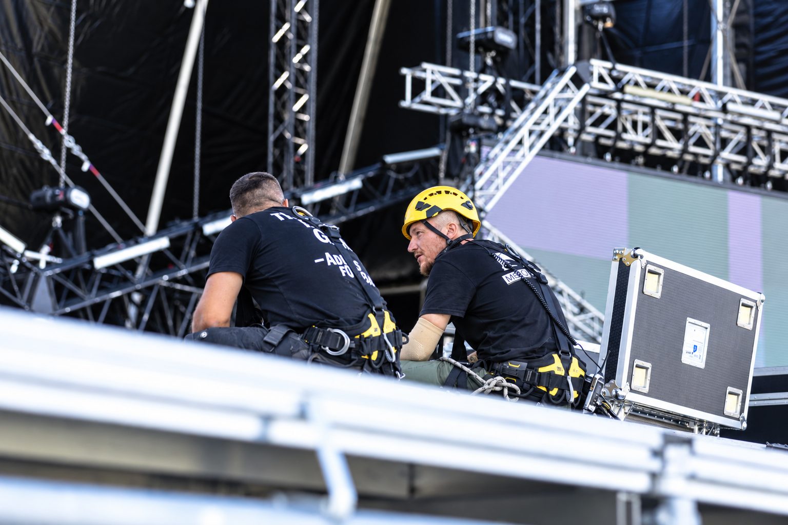Zwei Rigger mit Klettergurt und Helm arbeiten auf einer Bühnendachkonstruktion über Traversen und Kettenzügen