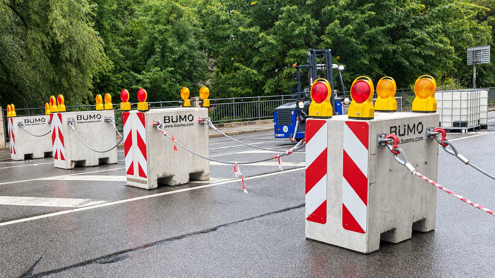 Perimeter Sicherung: Betonsperren mit Stahlseilen und gelben Warnleuchten sperren eine nasse Straße ab