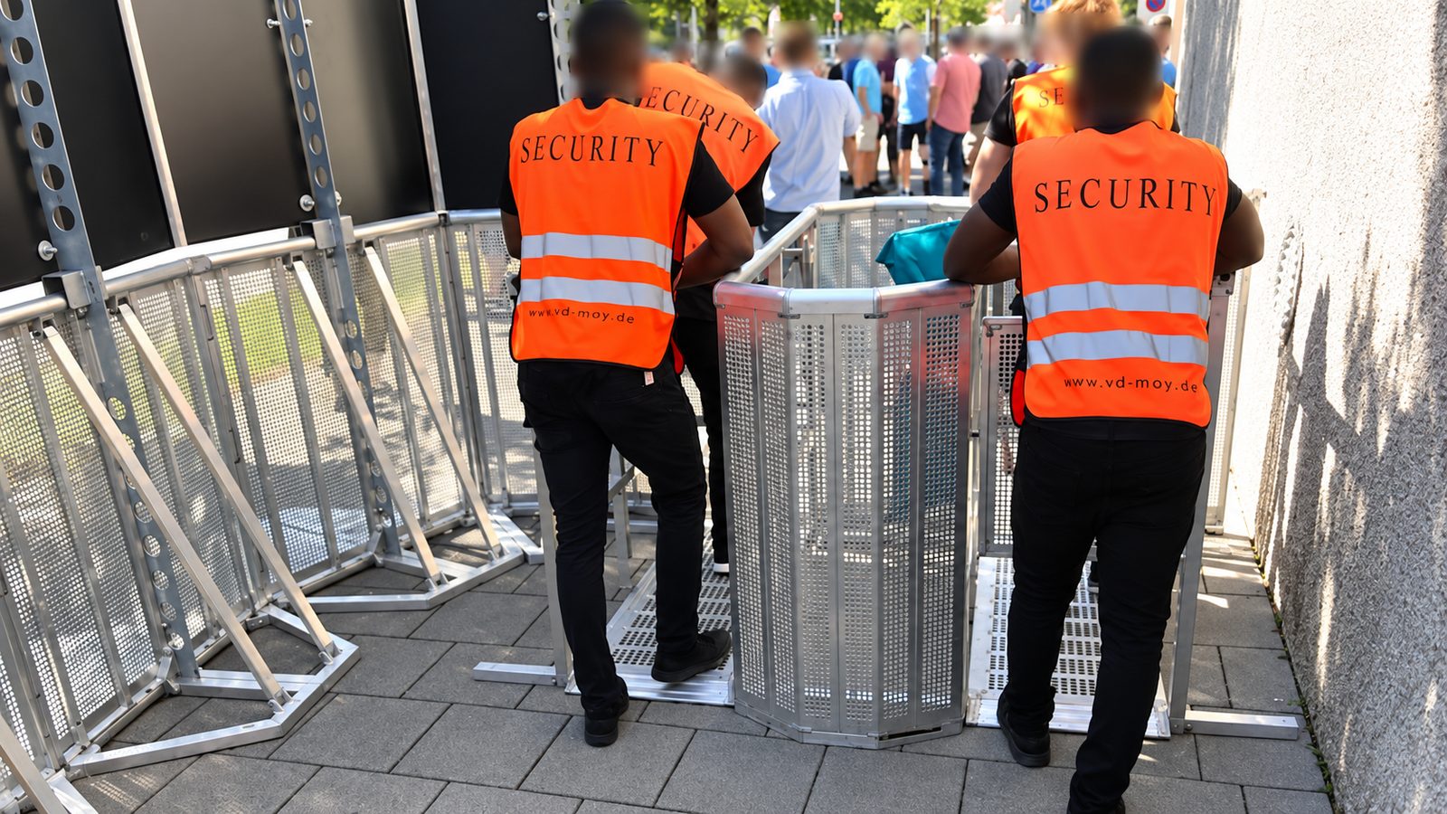 Zwei Ordner in orangefarbenen Warnwesten an einer Personenvereinzelungsanlage aus Aluminium