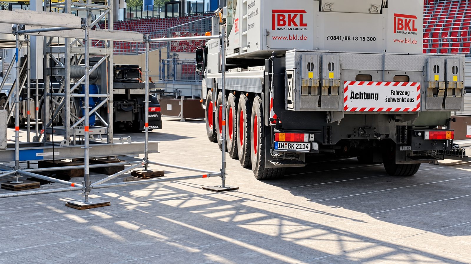 Gerüstfüße auf quadratischen Unterlagsplatten am Rand eines Betonplattenplatzes, dahinter ein Kranfahrzeug