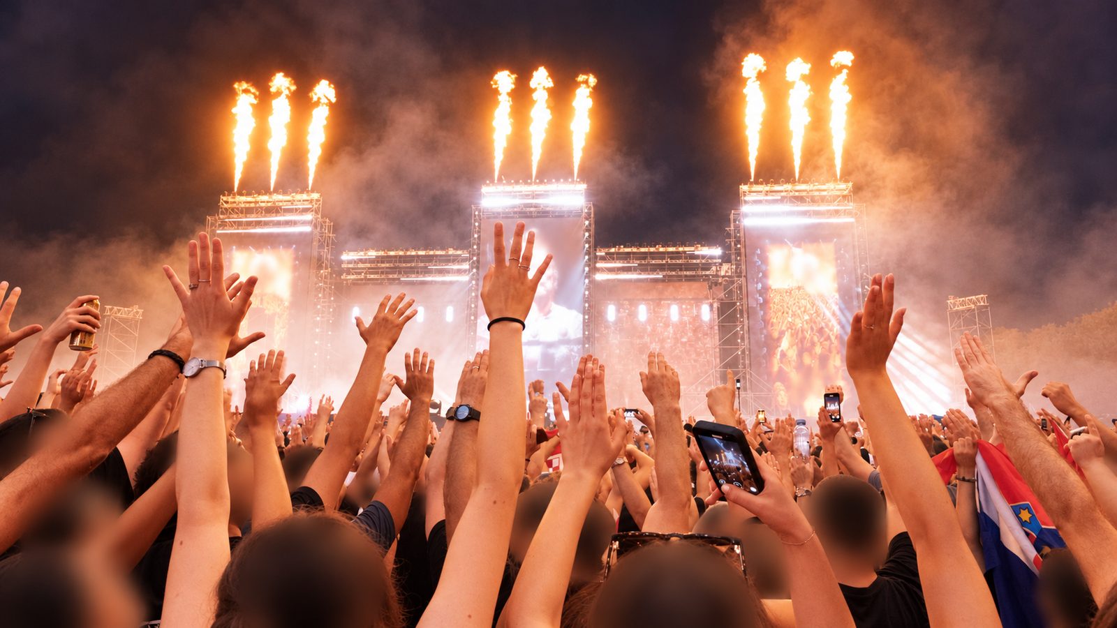 Dichtes Publikum bei Nacht mit erhobenen Händen vor einer Festivalbühne mit Flammeneffekten