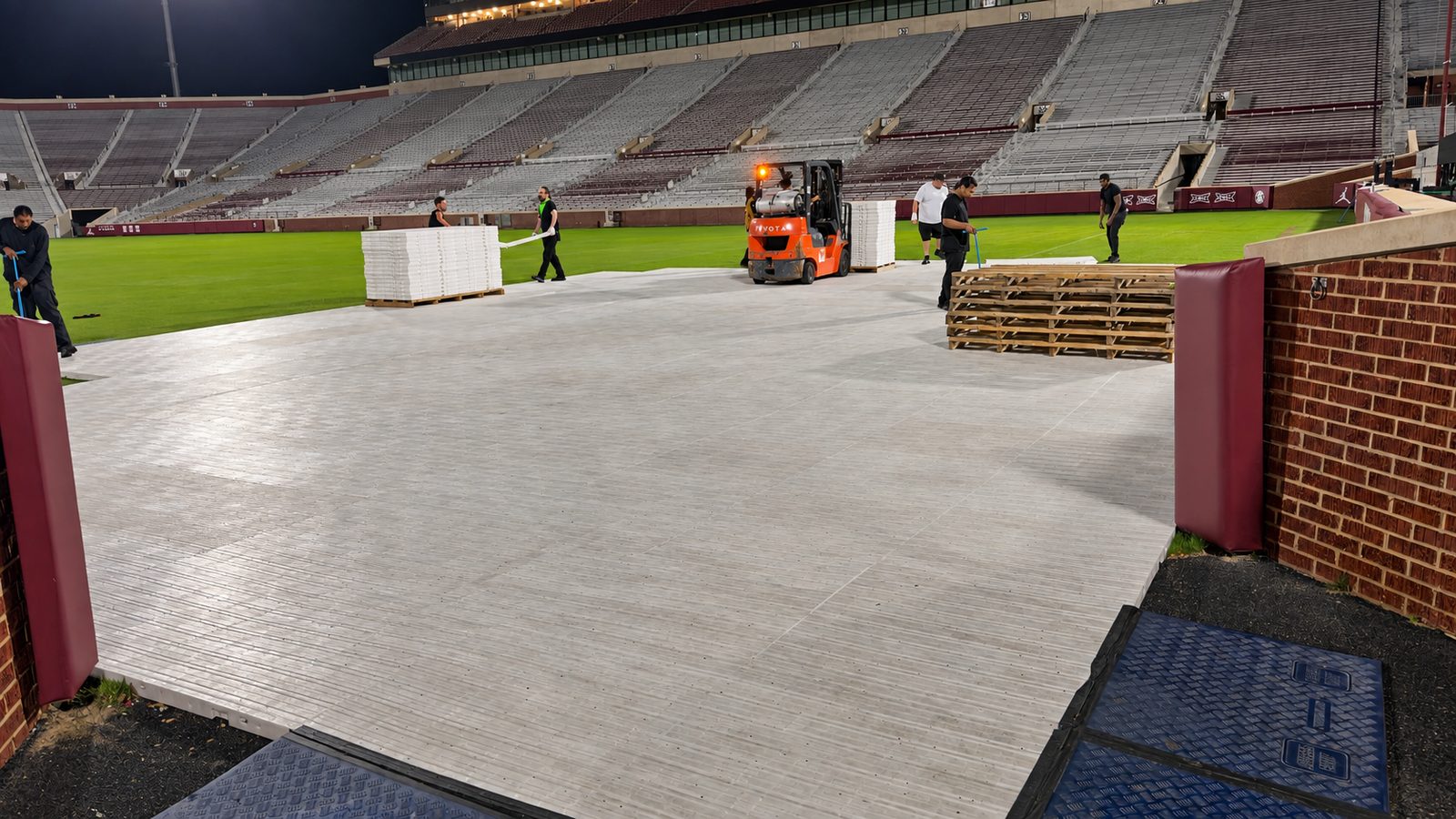 Nachtarbeit auf dem Spielfeld: fertig verlegte helle Plattenfläche, daneben offener Rasen und blaue Fahrplatten