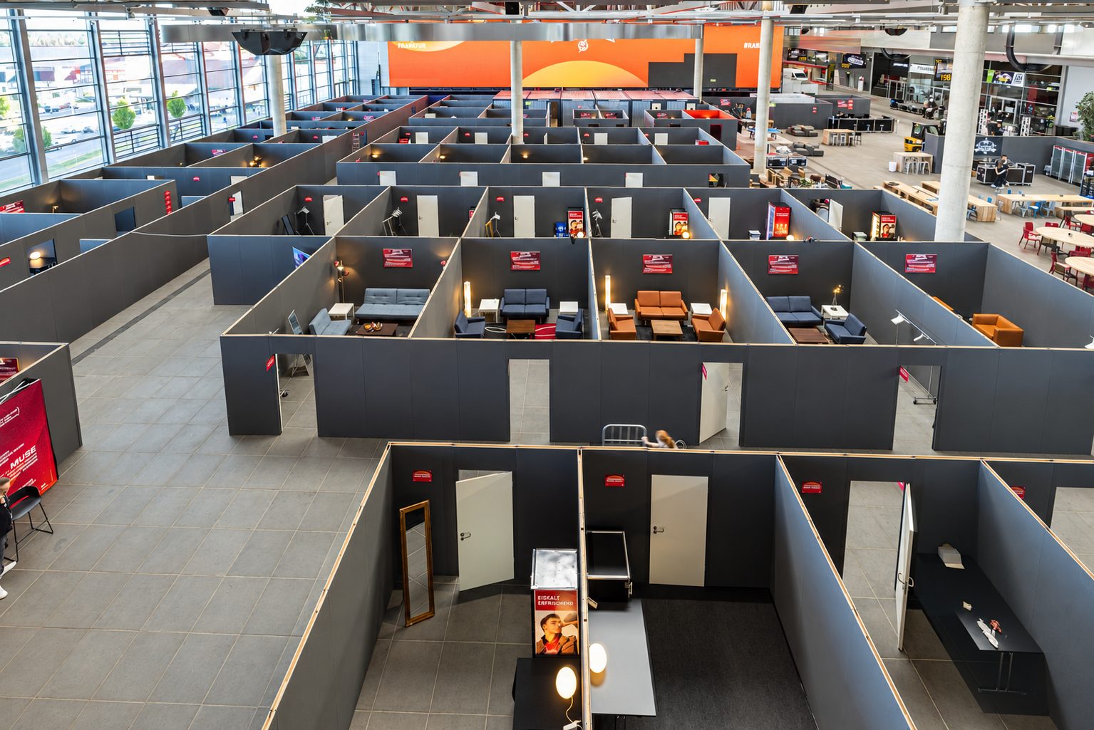 Raumsysteme aus grauen Trennwänden bilden in einer Halle ein Raster offener Kabinen mit Sitzmöbeln