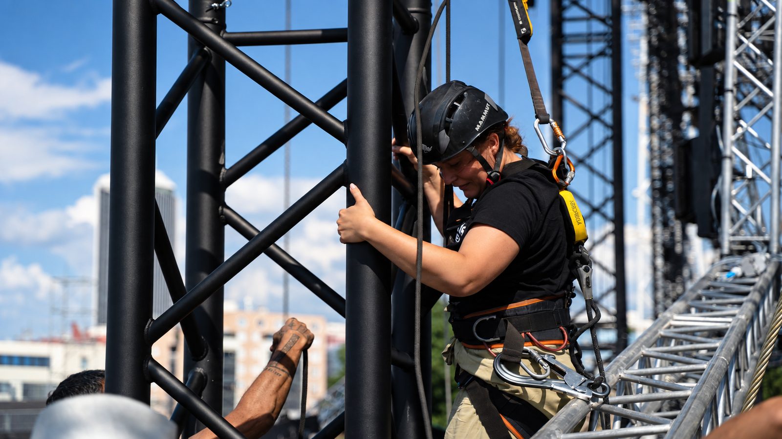 Rigging an der Bühnentraverse: Riggerin mit Helm und Klettergurt arbeitet an einem schwarzen Traversenturm
