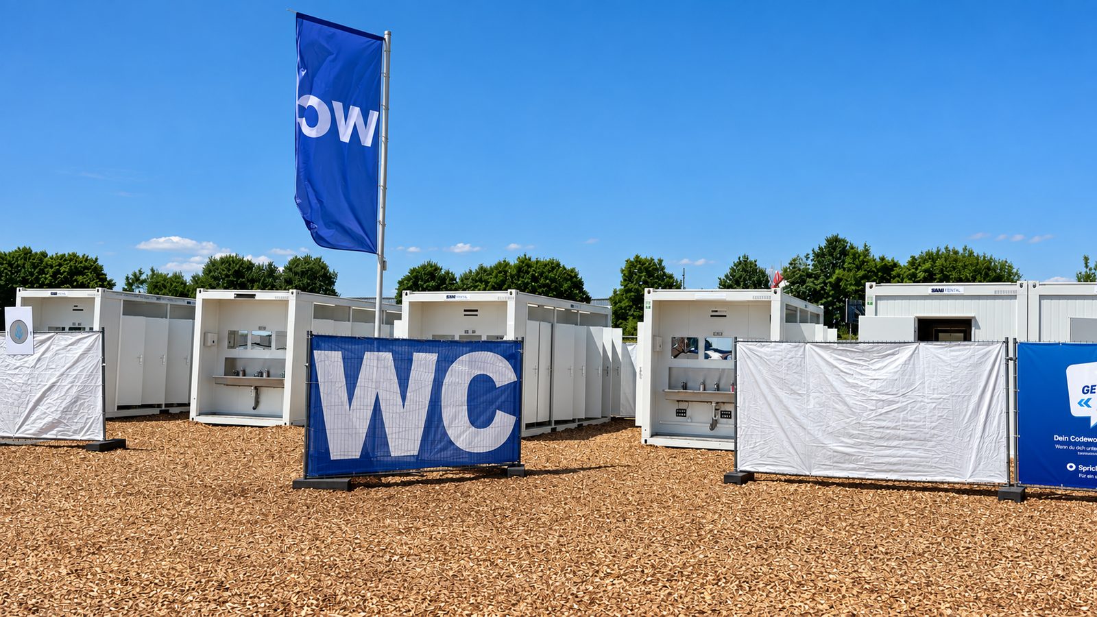 Reihe weißer WC-Container mit offenen Türen auf Hackschnitzelboden, davor ein blaues Banner mit der Aufschrift WC