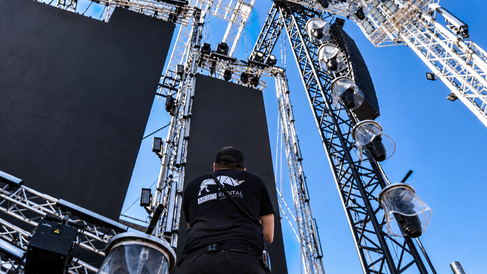 MOJO-Rental-Techniker in Untersicht vor Traversentürmen, dunklen LED-Wänden und einem Line-Array im Freien