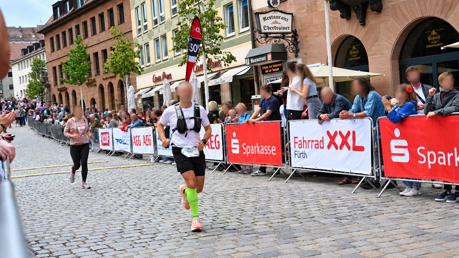 Sportevents in der Stadt: Läufer auf Kopfsteinpflaster, rechts bannerverkleidete Absperrgitter mit Zuschauern dahinter