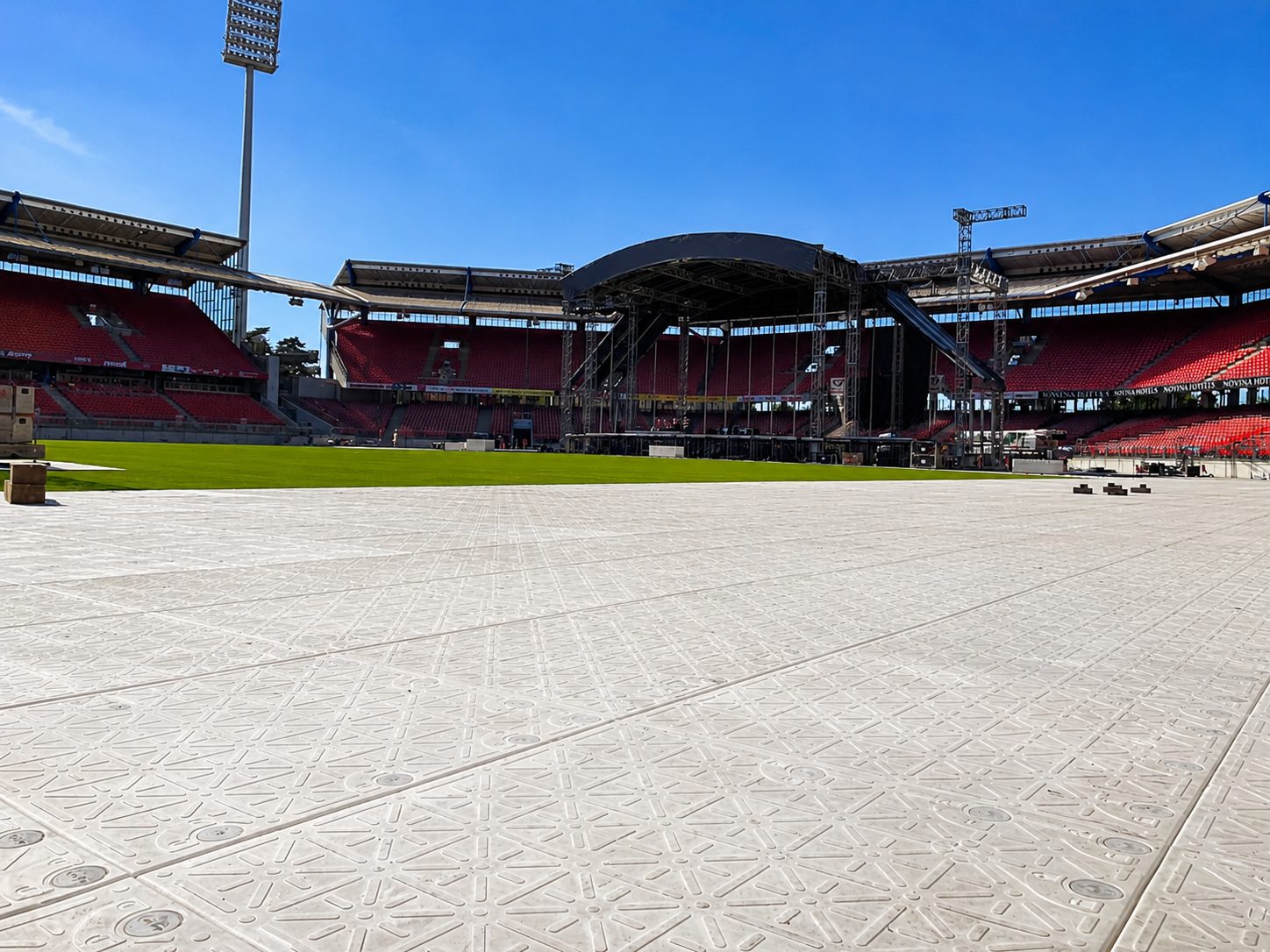 Stadion mit heller Bodenschutzplatten-Fläche im Vordergrund, dahinter offener Rasen und eine überdachte Bühne
