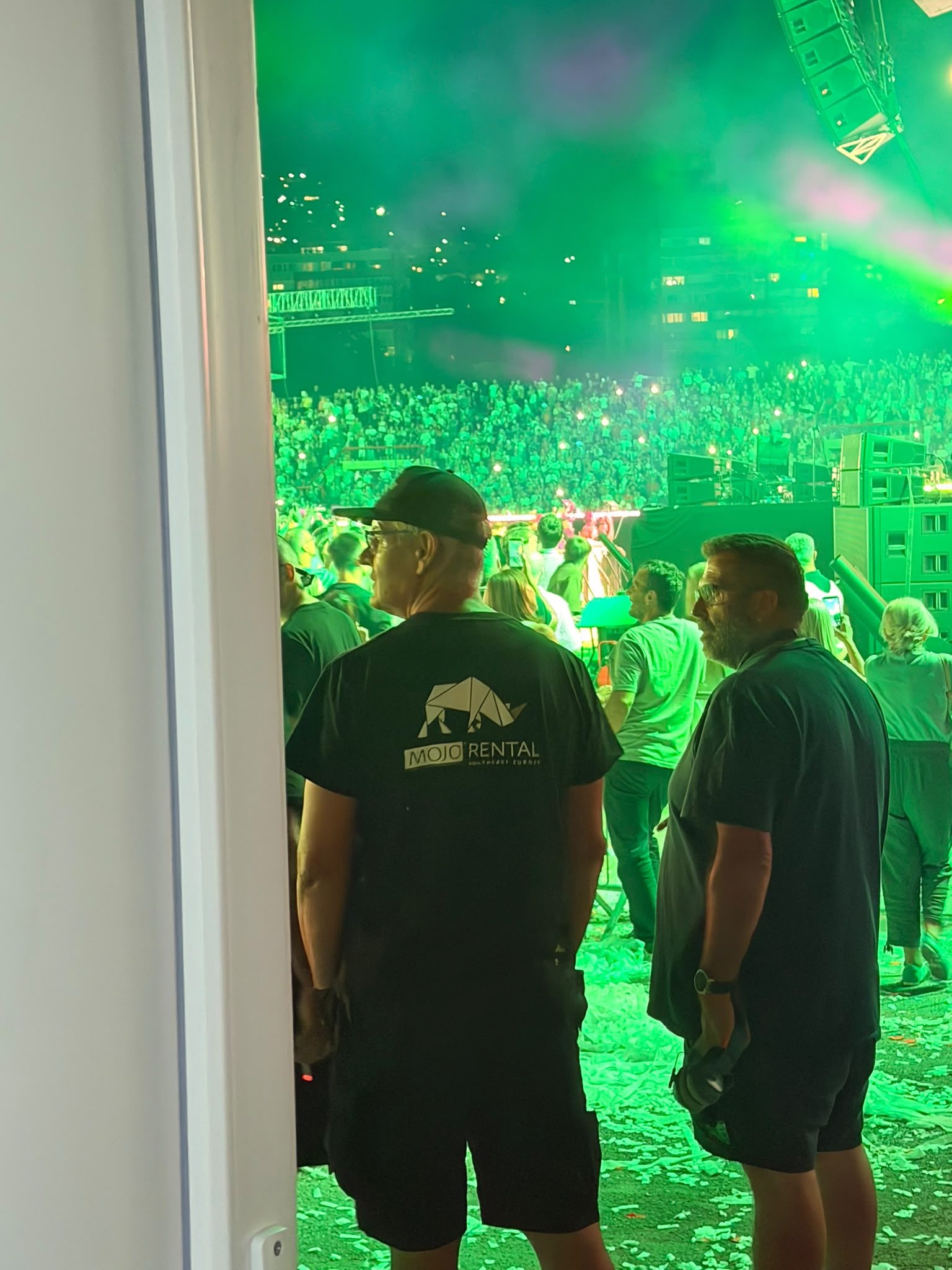 Blick aus einer Türöffnung auf ein Open-Air-Konzert bei Nacht, grünes Bühnenlicht über dichtem Publikum