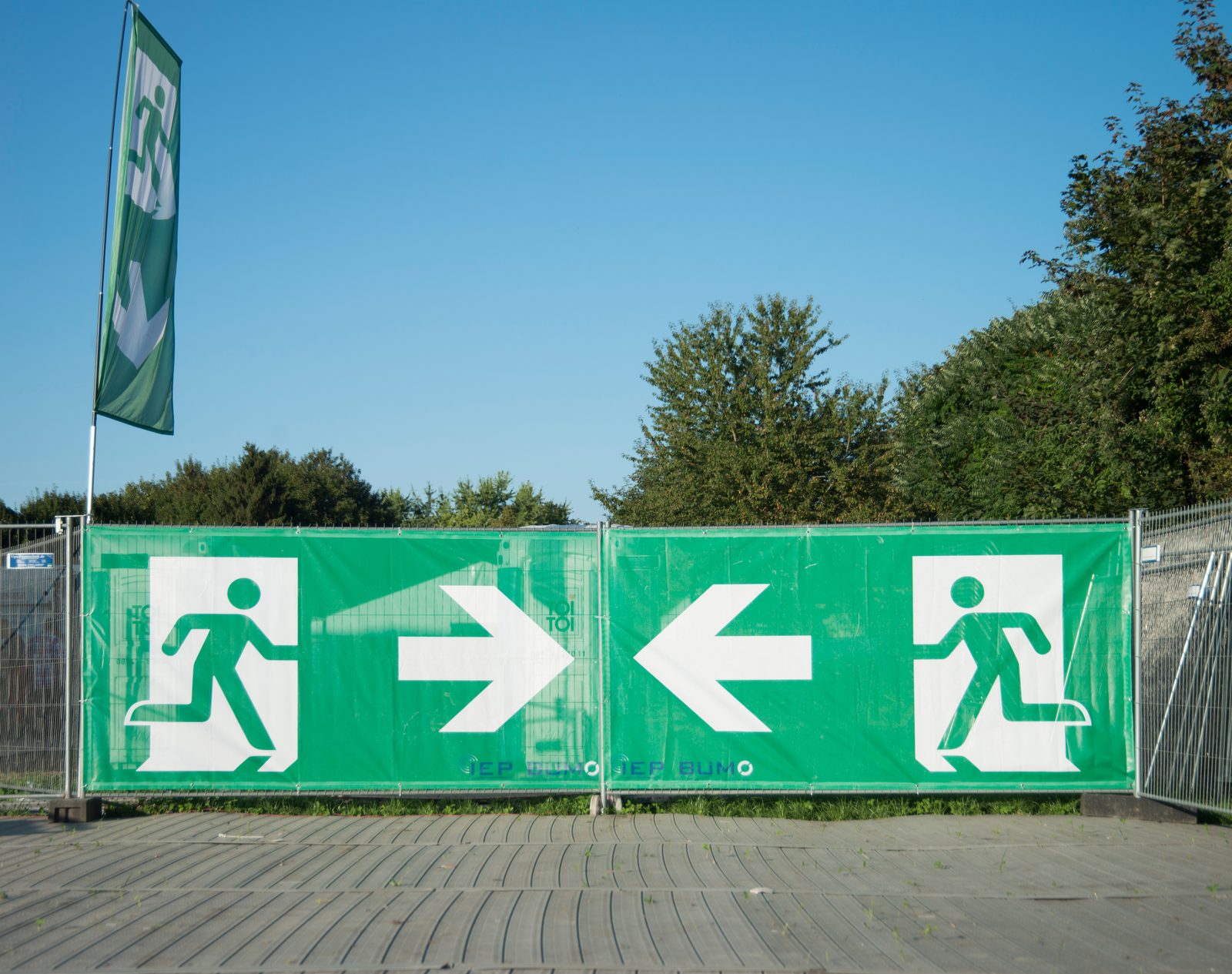 Bauzaun mit grünen Fluchtwegbannern, weiße Piktogramme und Pfeile weisen nach links und rechts