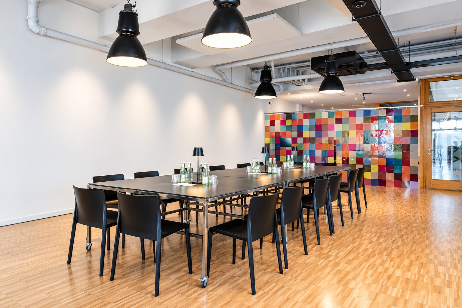 Besprechungsraum mit schwarzem Rolltisch, Industrieleuchten und einer Wand aus bunten Quadraten.