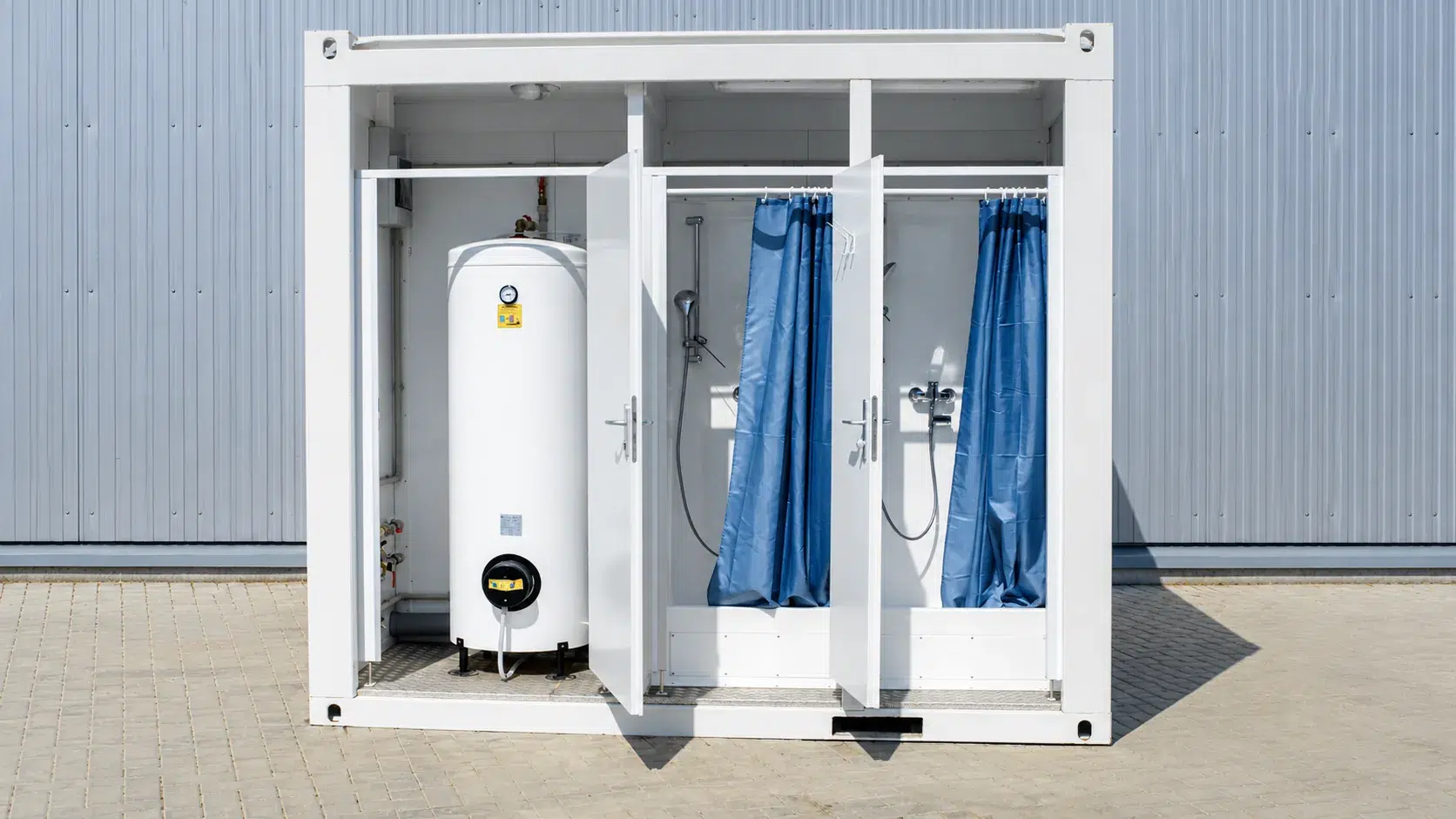 Geöffneter weißer Container mit Warmwasserboiler und zwei Duschkabinen mit blauen Vorhängen vor einer Hallenwand