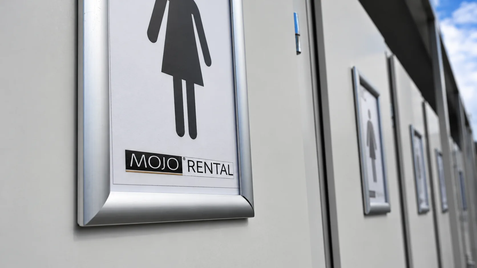 Nahaufnahme eines Damen-Piktogramms mit MOJO-Rental-Logo an der Tür eines weißen Toilettenwagens