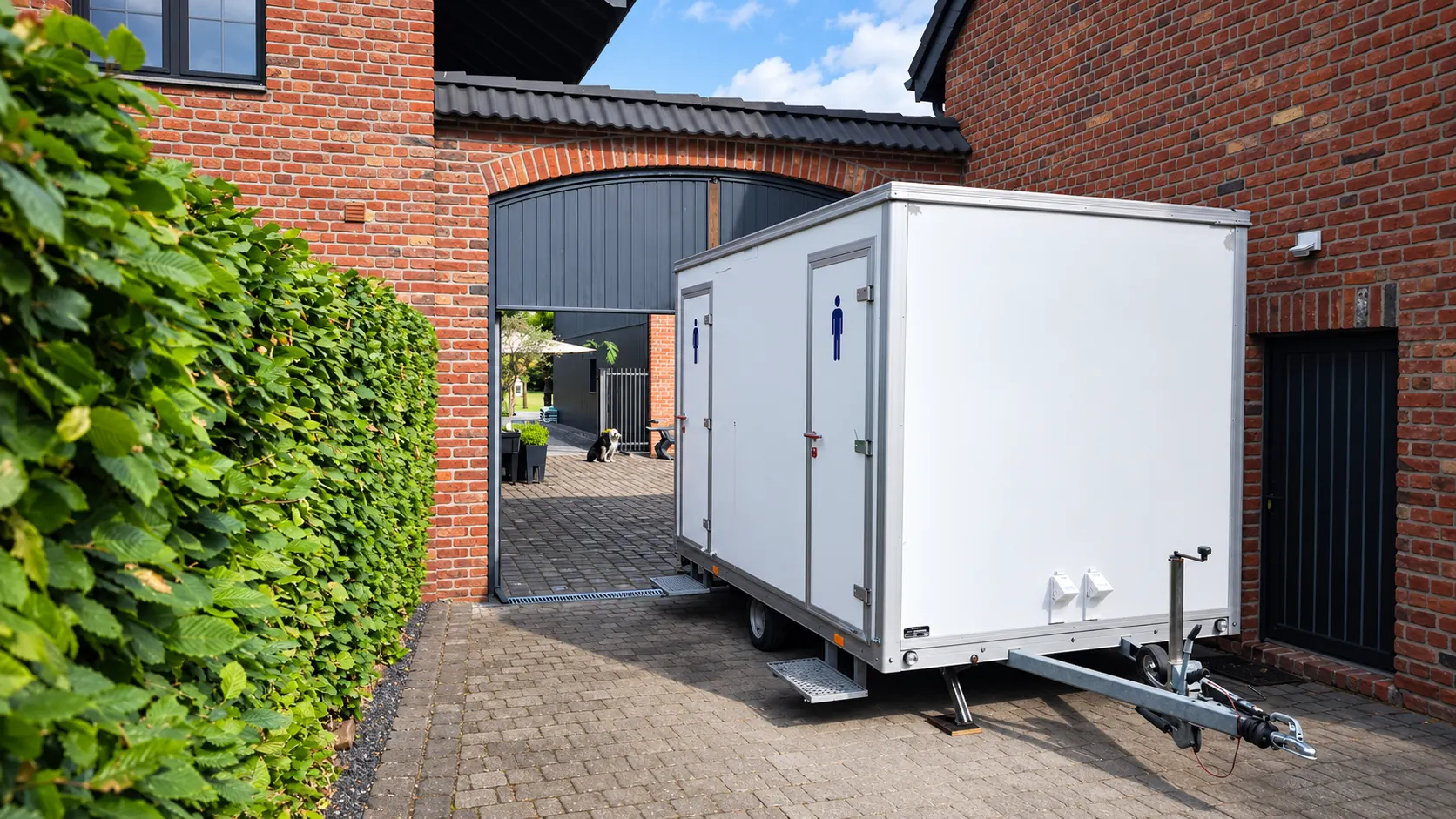 Kompakter weißer Toilettenwagen mit Damen- und Herrentür im gepflasterten Innenhof eines Backsteingebäudes