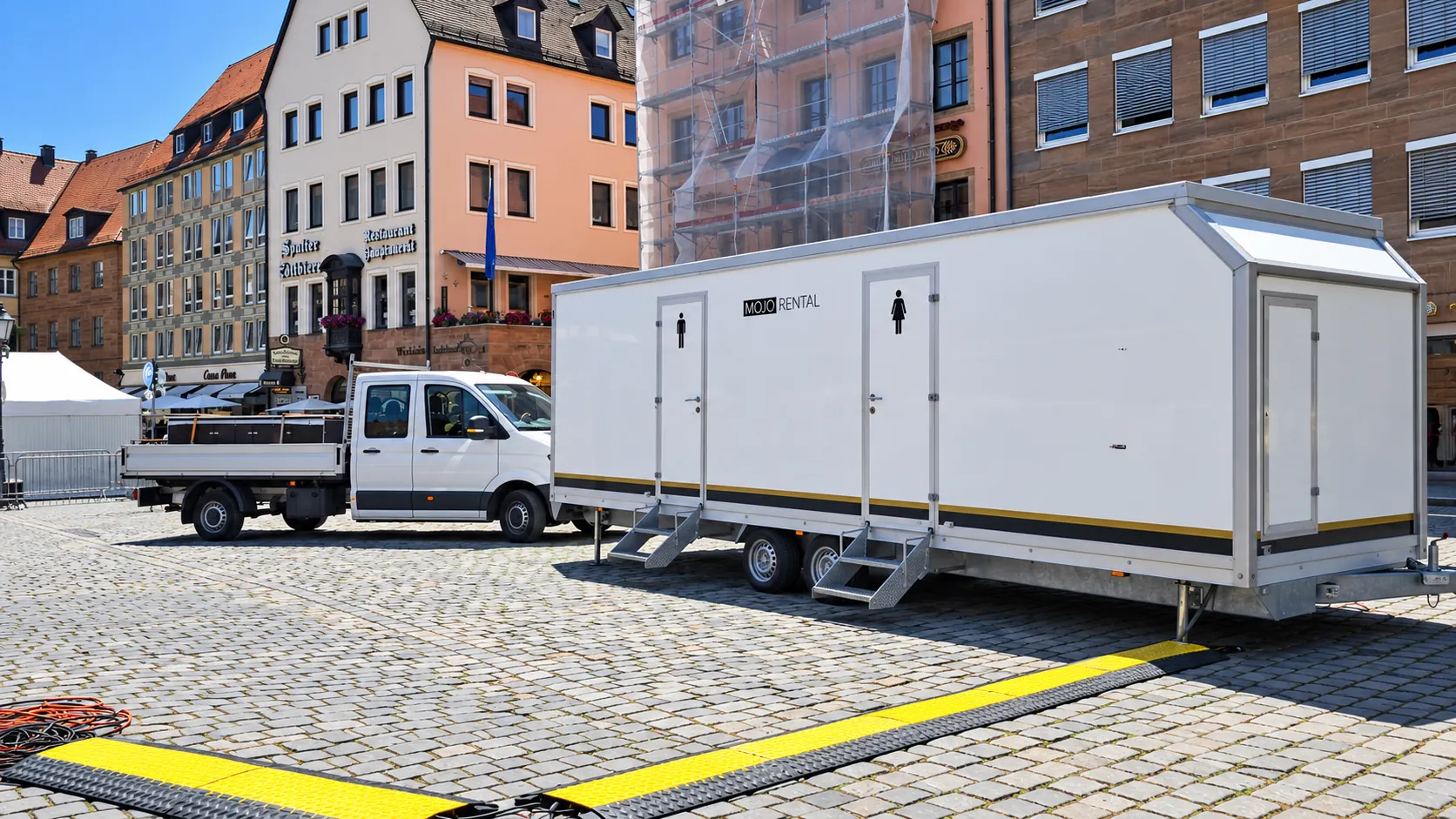 Weißer Toilettenwagen mit Damen- und Herrentür auf einem Pflasterplatz in der Altstadt, daneben ein Pritschenwagen